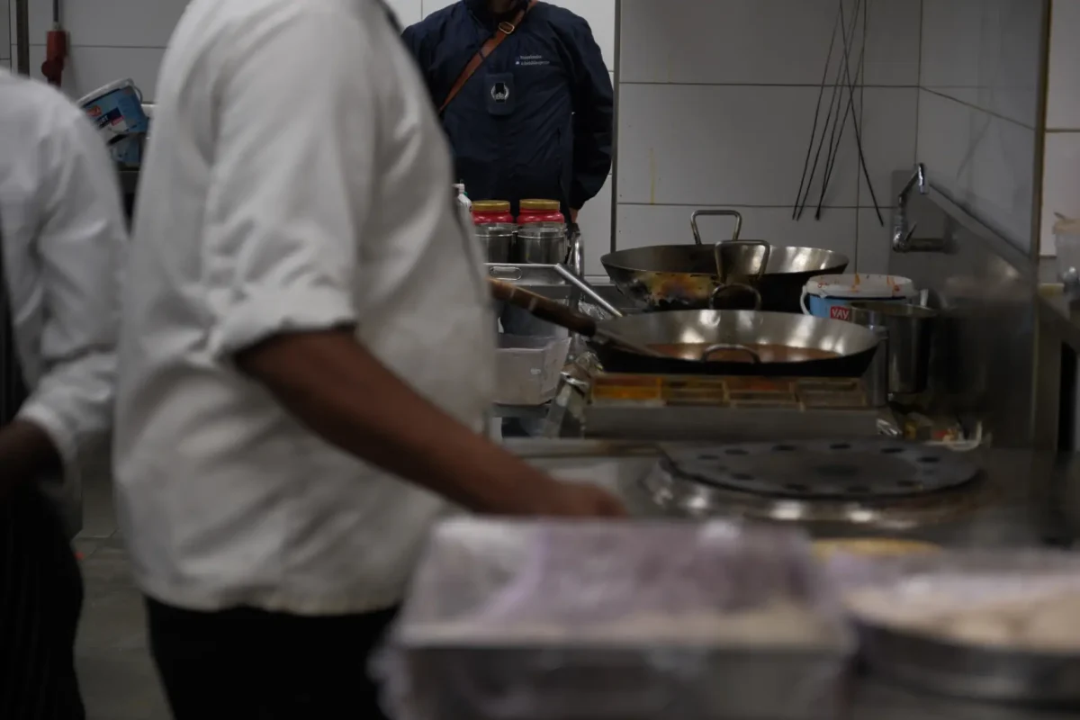 In einer Restaurantküche ist ein Polizist im Hintergrund zu sehen, während ein Koch im Vordergrund arbeitet. Auf dem Herd stehen mehrere Pfannen.