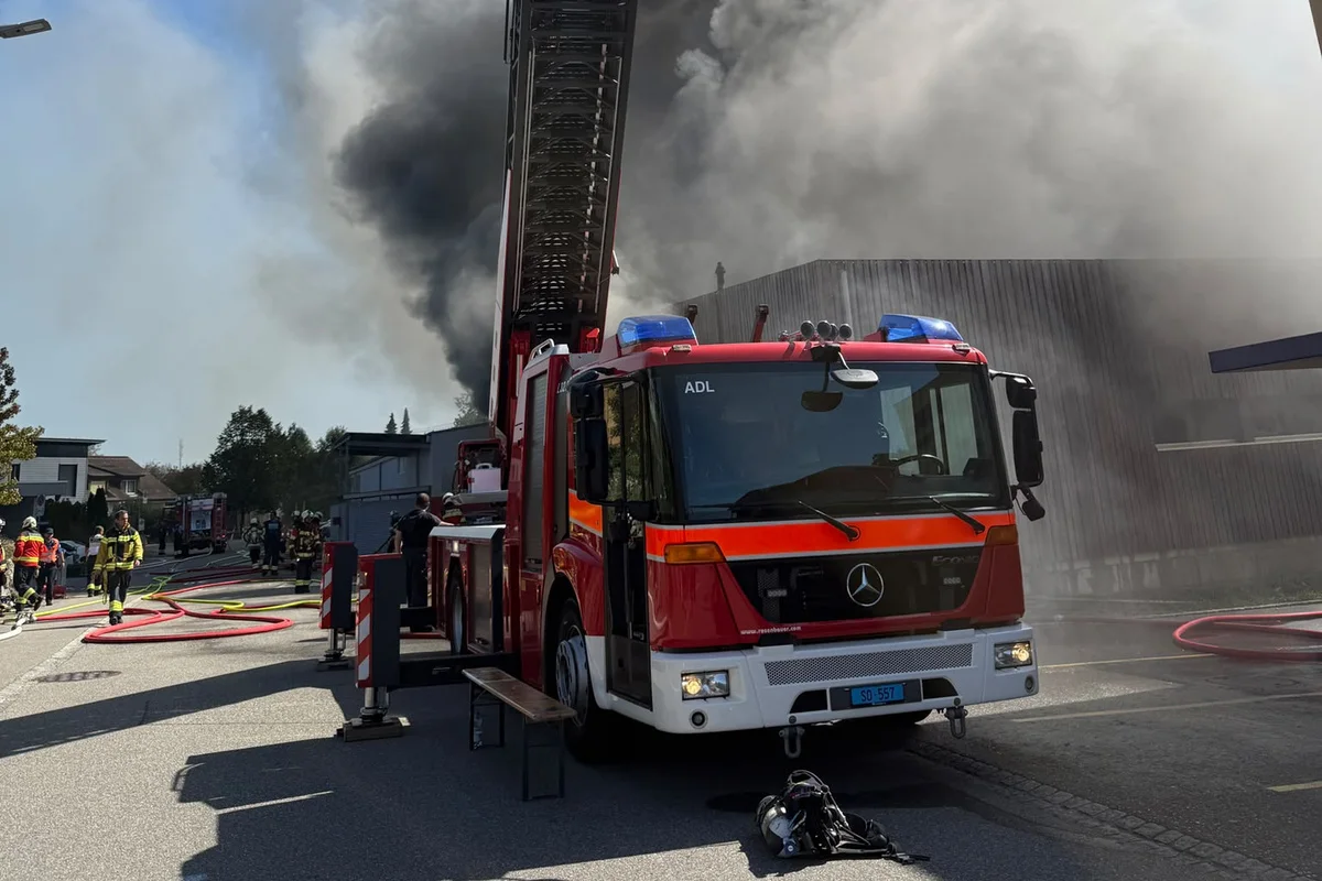 Ein rotes Feuerwehrfahrzeug mit ausgefahrener Drehleiter steht vor einem stark rauchenden Gebäude. Feuerwehrleute stehen in der Nähe mit Schläuchen bereit.