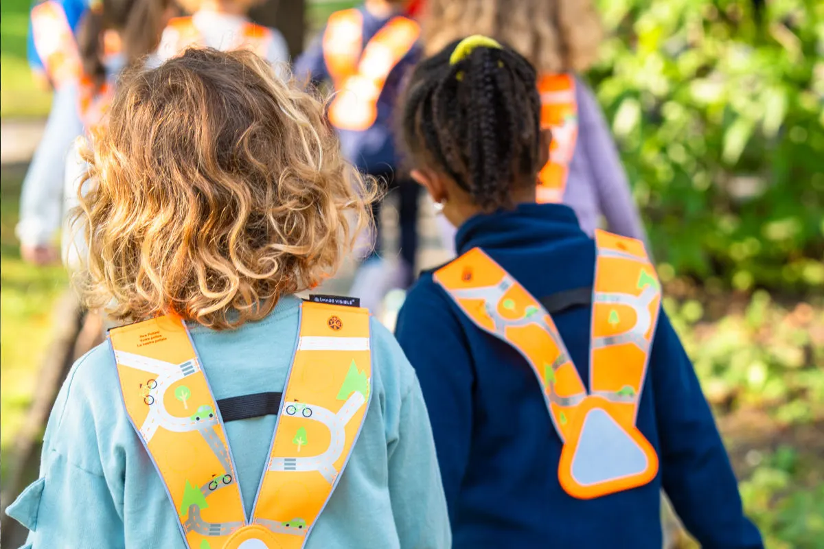 Mehrere Kinder mit orangefarbenen Warnwesten sind von hinten zu sehen, während sie im Freien laufen.