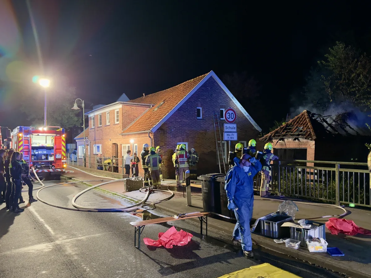 Mehrere Feuerwehrleute sind im Einsatz neben einem brennenden Gebäude, sichtbare Rauchentwicklung bei Nacht.