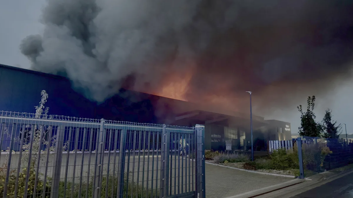 Ein Industriegebäude steht in Flammen; dichter Rauch steigt auf. Das Gebäude ist von einem Sicherheitszaun umgeben.