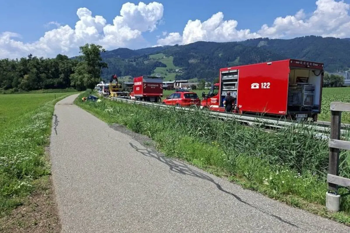 Mehrere Feuerwehrfahrzeuge und ein rotes Auto sind auf einer Landstraße im Grünen zu sehen. Im Hintergrund erheben sich bewaldete Hügel unter einem klaren Himmel.