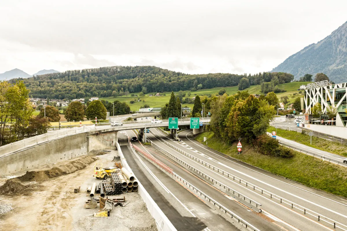 Das Bild zeigt eine Autobahnbaustelle mit einer Brücke im Vordergrund. Im Hintergrund sind Bäume und eine hügelige Landschaft sichtbar.