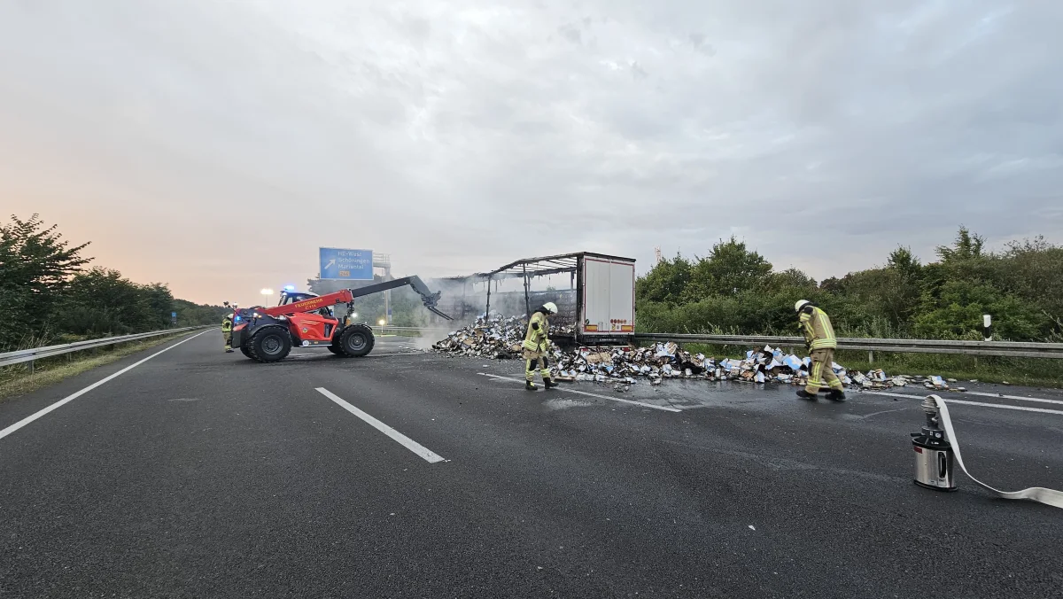 Auf der Autobahn sind Feuerwehrleute dabei, die Überreste einer verbrannten LKW-Ladung zu beseitigen. Ein Feuerwehrfahrzeug unterstützt den Einsatz.