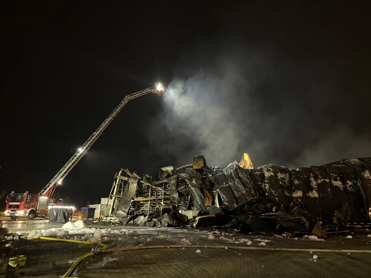 Ein Feuerwehrwagen mit einem hochgefahrenen Lichtträger beleuchtet die Überreste eines ausgebrannten Lagerhauses. Rauch steigt noch von den Trümmern in die Nachtluft.