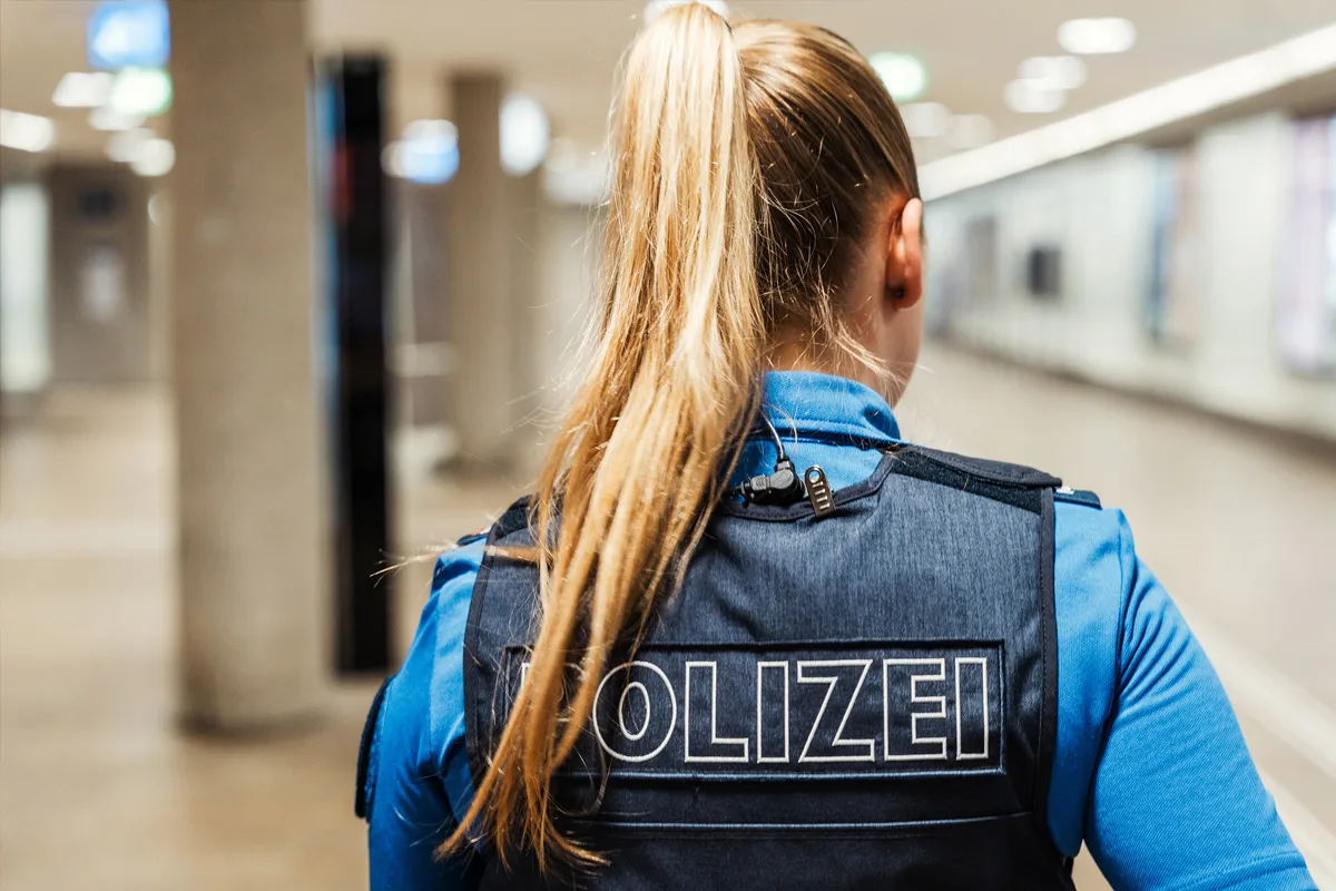 Man sieht die Rückansicht einer Polizistin mit blauer Uniform und Headset, die einen Flur überwacht. Der Flur wirkt modern und gut beleuchtet.