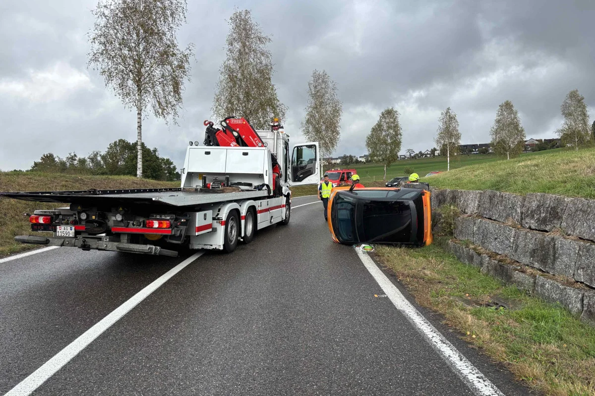 Ein orangenes Auto liegt auf der Seite neben einer nassen Landstraße. Ein Abschleppfahrzeug und zwei Personen in Warnwesten sind vor Ort.
