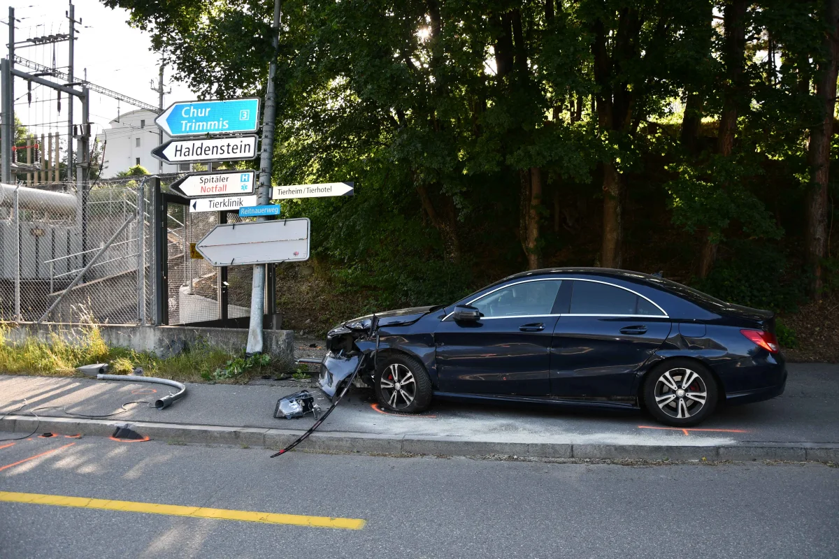Ein schwarzes Auto mit Frontschaden steht am Straßenrand, umgeben von Bäumen und verschiedenen Verkehrsschildern.