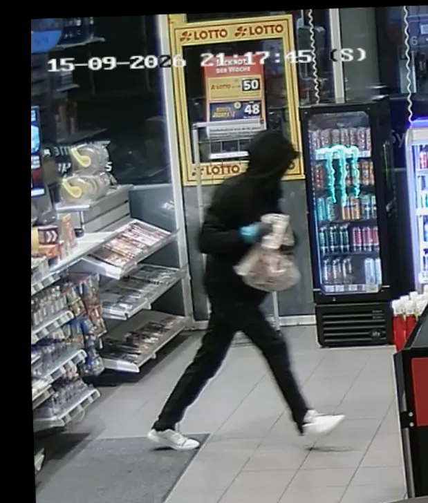 Eine vermummte Person in dunkler Kleidung und blauen Handschuhen hält eine Tasche und läuft durch einen Laden, gefilmt von einer Überwachungskamera.