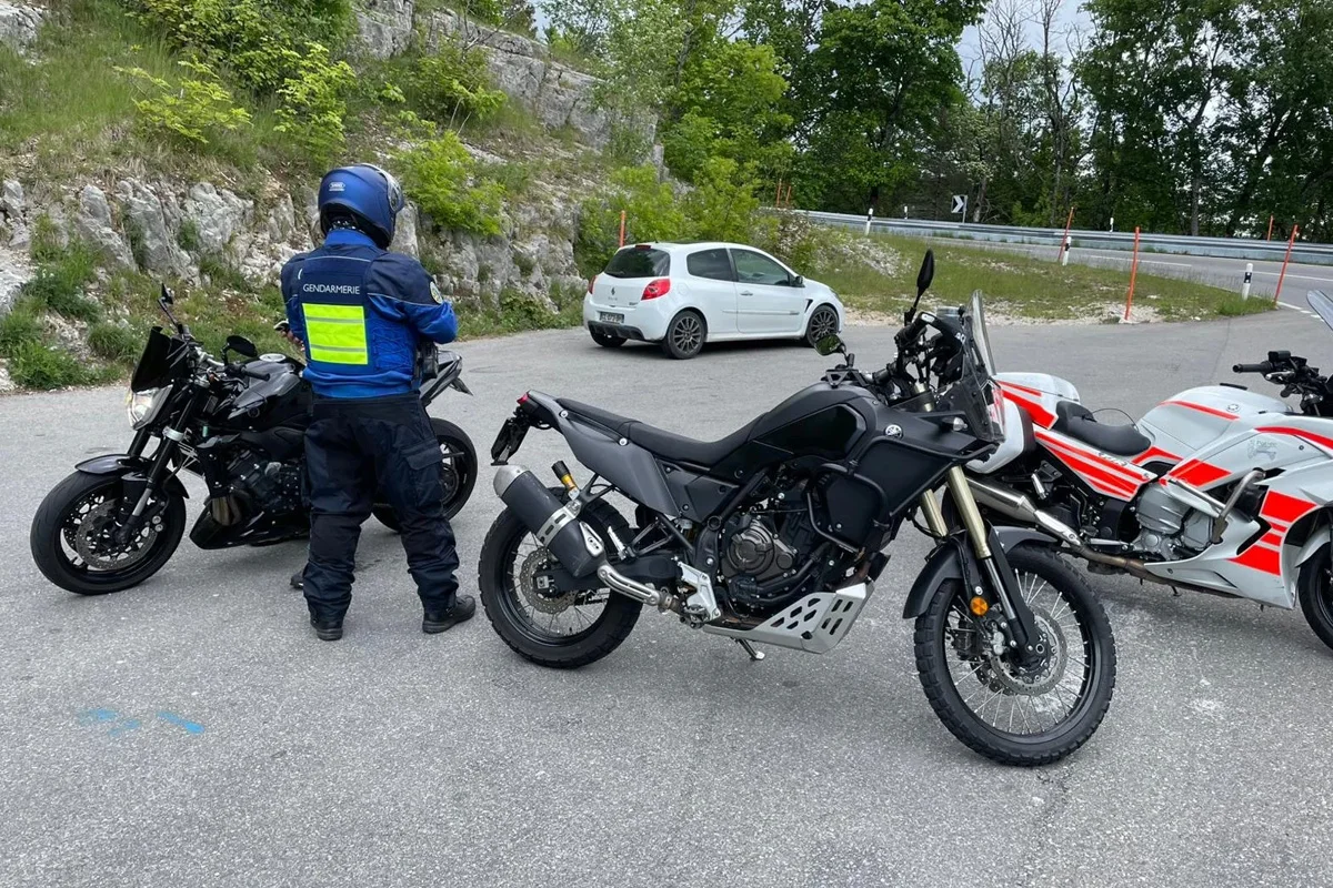 Ein Polizist in blauer Uniform steht auf einer Straße neben drei abgestellten Motorrädern und einem Auto im Hintergrund. Es ist eine landwirtschaftliche Umgebung mit Bäumen und Felsen sichtbar.