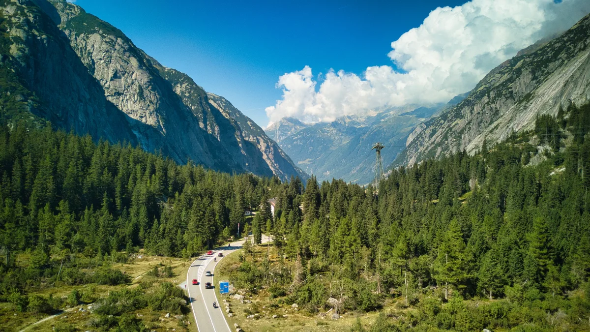 Eine Straße windet sich durch ein alpines Tal. Auf der Straße sind mehrere Fahrzeuge zu sehen, umgeben von Bergen und dichten Wäldern unter einem blauen Himmel.