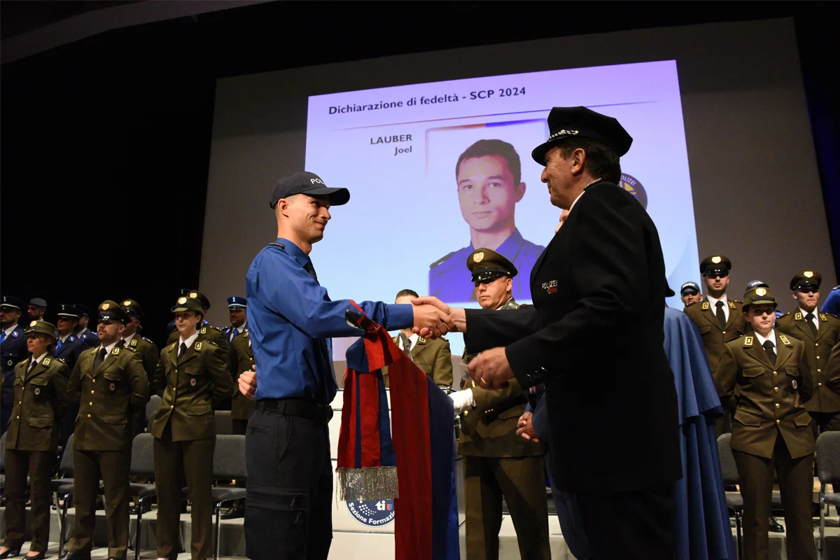 Eine Person in blauer Polizeiuniform schüttelt während einer Zeremonie die Hand eines anderen in dunkler Uniform. Im Hintergrund sind weitere Polizisten in Uniform zu sehen und eine Projektion auf einer Leinwand.