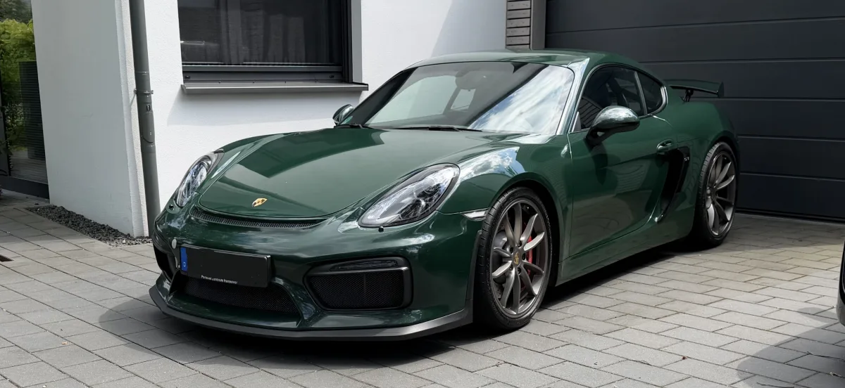 Ein dunkelgrüner Porsche ist auf einer gepflasterten Einfahrt vor einem modernen weißen Gebäude geparkt. Im Hintergrund ist ein Fenster und ein Garagentor sichtbar.