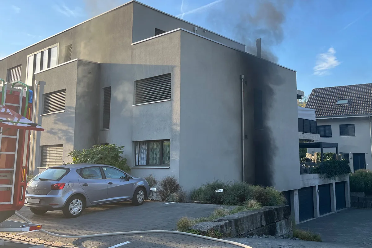 Ein graues Wohnhaus mit dunklem Rauch, der an der Fassade aufsteigt. Vor dem Haus parkt ein silbernes Auto.