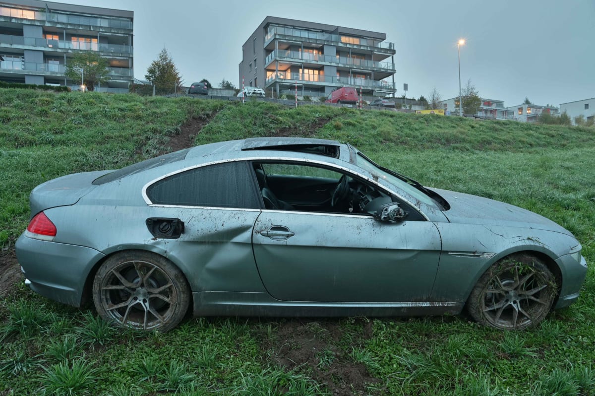 Das Bild zeigt ein silberfarbenes Auto mit sichtbaren Schäden auf einer Wiese. Im Hintergrund sind moderne Wohnhäuser zu sehen.