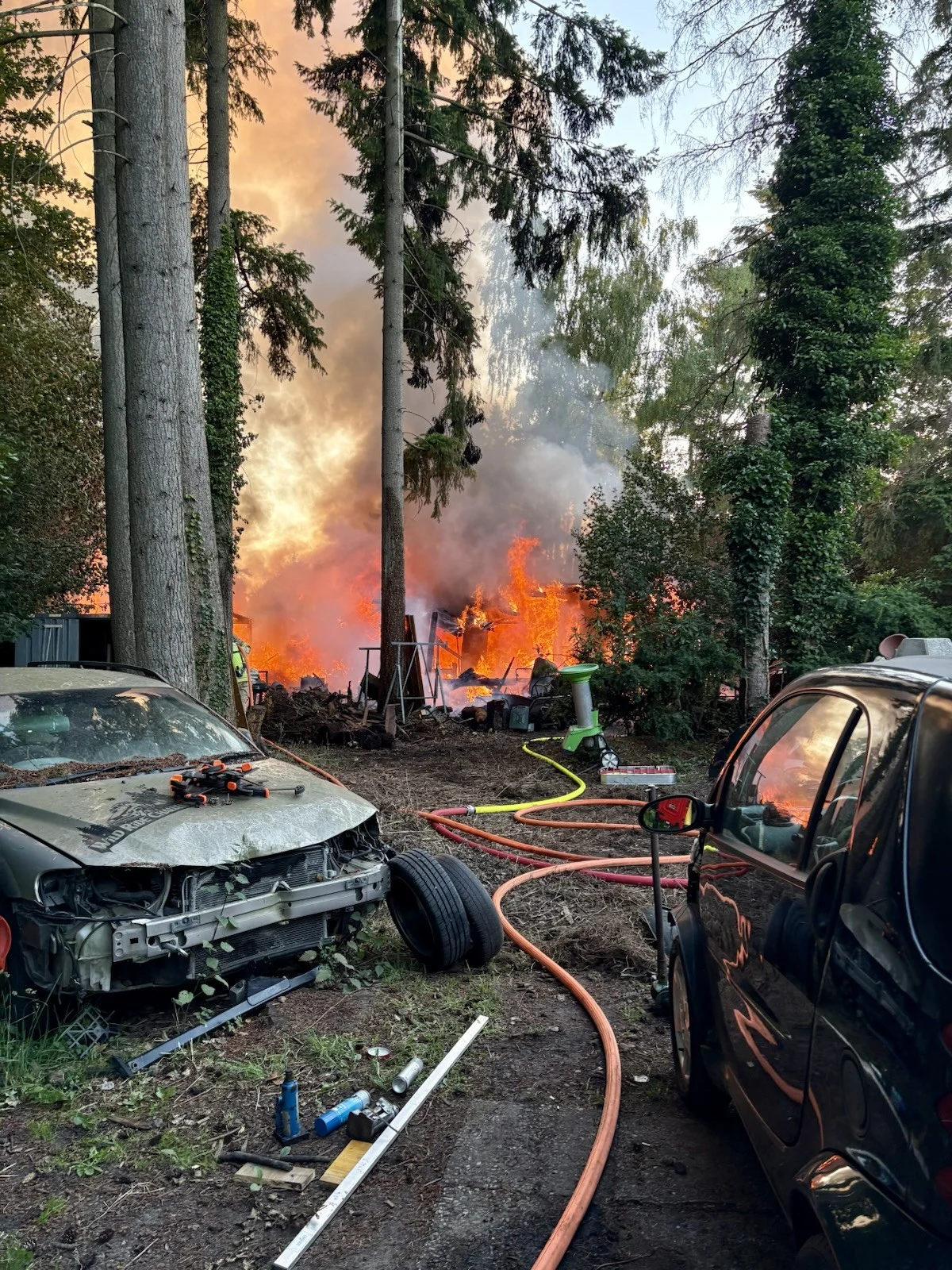 Ein intensives Feuer lodert in einem Waldgebiet, Flammen und Rauch sind deutlich sichtbar. Im Vordergrund stehen beschädigte Autos.