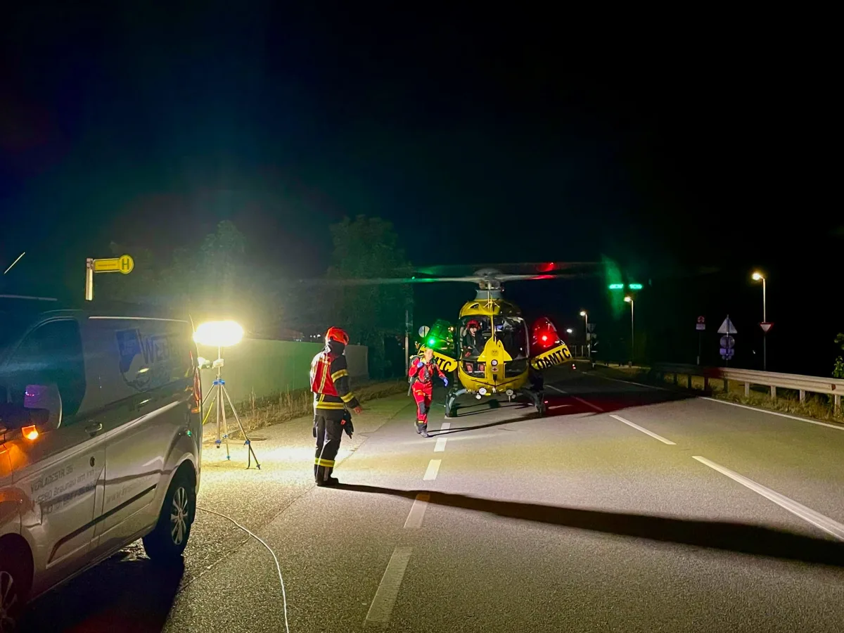 Ein gelber Rettungshubschrauber steht nachts auf einer Straße. Einsatzkräfte in Uniform sind umgeben von Scheinwerfern.