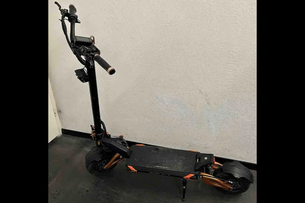 Ein schwarzer E-Scooter steht auf einem dunklen Boden, gelehnt an eine weiße Wand. Er zeigt breite Reifen und ein Display auf dem Lenker.