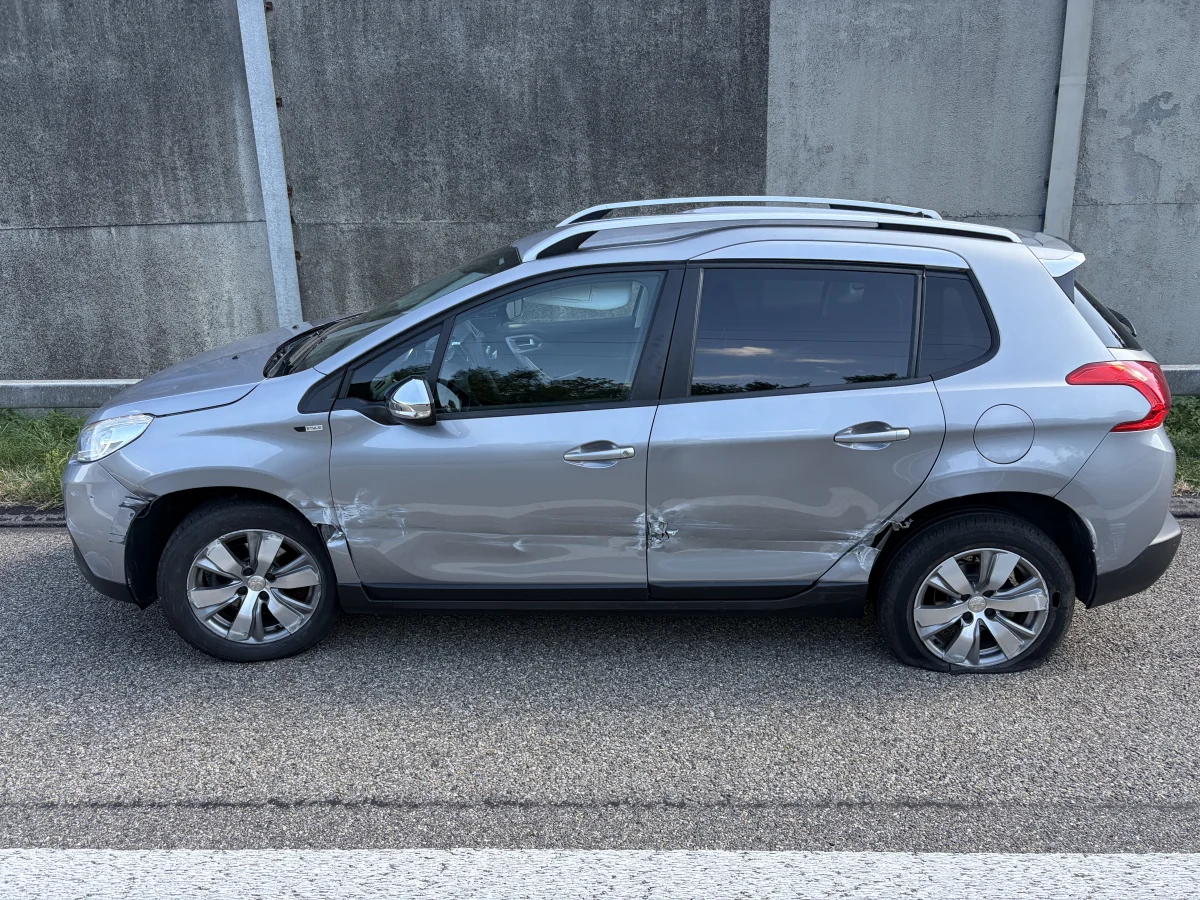 Ein silbernes Auto mit sichtbaren Beschädigungen an der linken Seite steht am Straßenrand vor einer Betonwand.