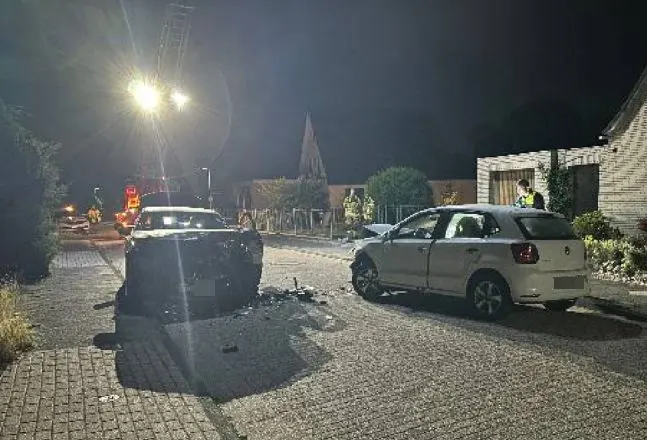 Bei Nacht sind zwei beschädigte Fahrzeuge auf einer gepflasterten Straße zu sehen. Ein Auto hat sichtbare Frontschäden, während ein Polizist die Unfallstelle untersucht.