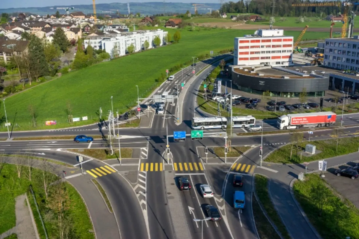 Eine Luftaufnahme zeigt eine stark frequentierte Kreuzung mit Autos und einem Bus in einem Vorstadtbereich. Mehrere Straßen führen von und zur Kreuzung, umgeben von Wohnhäusern und Grünflächen.