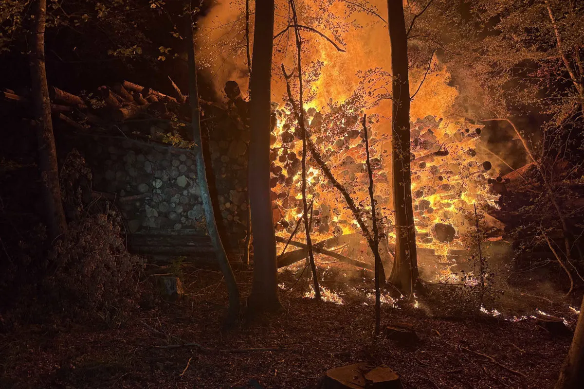 Ein großer Stapel von Holzstämmen brennt mit starken Flammen mitten in einem nächtlichen Wald. Die Umgebung wirkt dunkel und die Flammen dominieren die Szene.