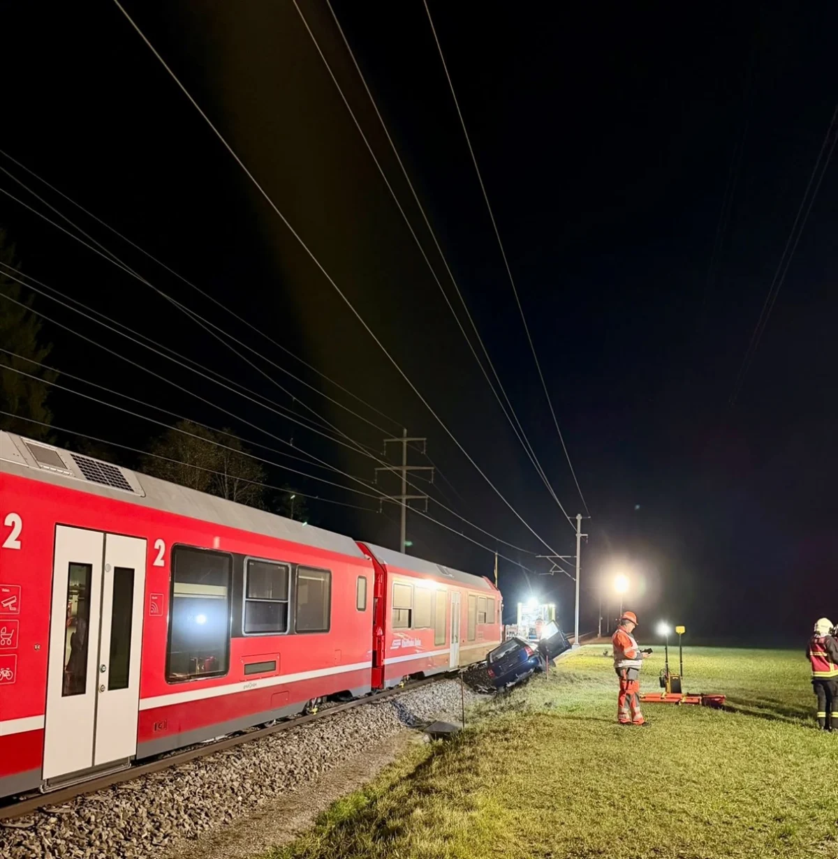 Ein roter Zug steht auf einem Bahngleis. Daneben sind Scheinwerfer, Einsatzkräfte und ein verunfalltes Fahrzeug zu erkennen.