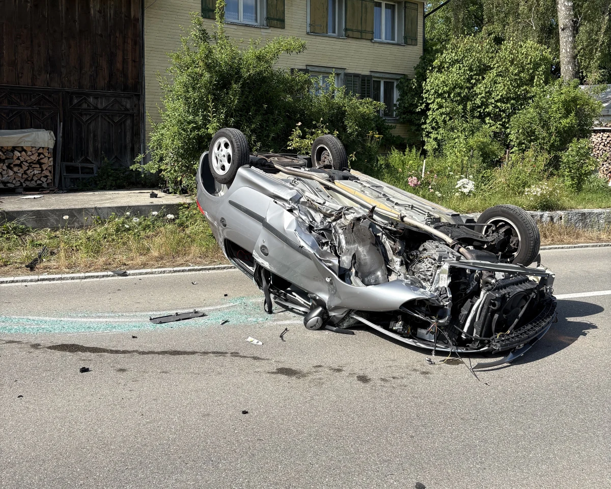 Ein silbernes Auto liegt stark beschädigt auf dem Dach auf einer Landstraße. Umgeben von Bäumen und einem Haus im Hintergrund.