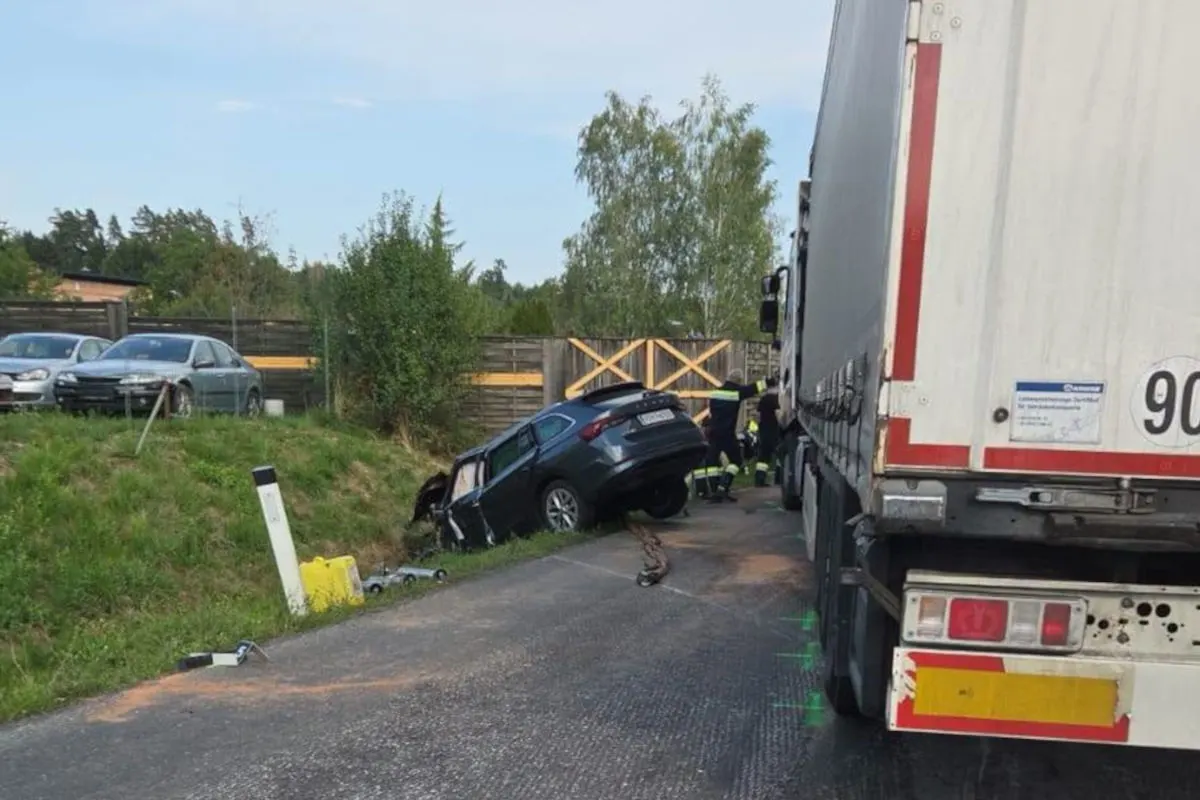 Ein Auto ist im Graben neben einer Straße zu sehen, während rechts ein Lastwagen steht. Männer in Uniformen stehen daneben.