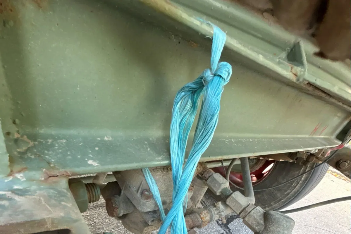 Das Bild zeigt ein blaues Seil, das improvisiert um eine Metallstange eines Fahrzeugs gebunden ist. Man sieht den rostigen Rahmen und Teile des Anhängerreifens im Hintergrund.