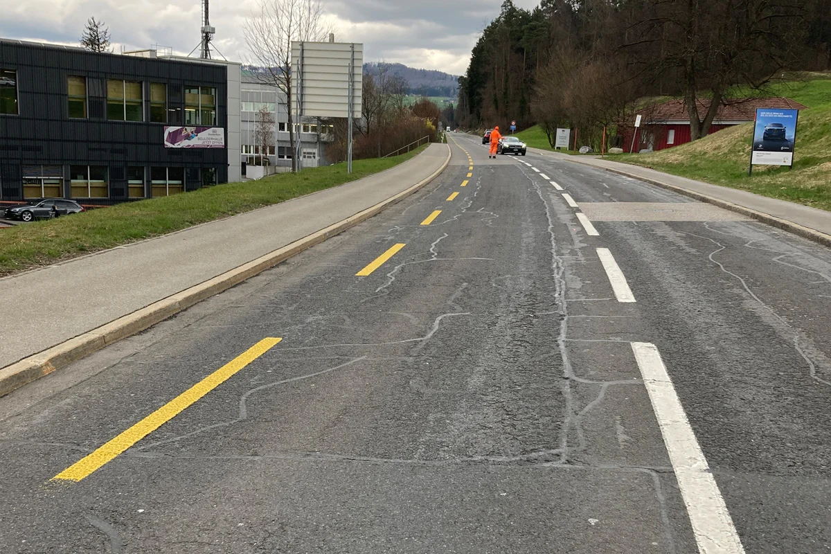Eine Straße mit gelben Markierungen, ein Arbeiter in Warnkleidung steht neben einem Fahrzeug. Rechts sind Bäume zu sehen.