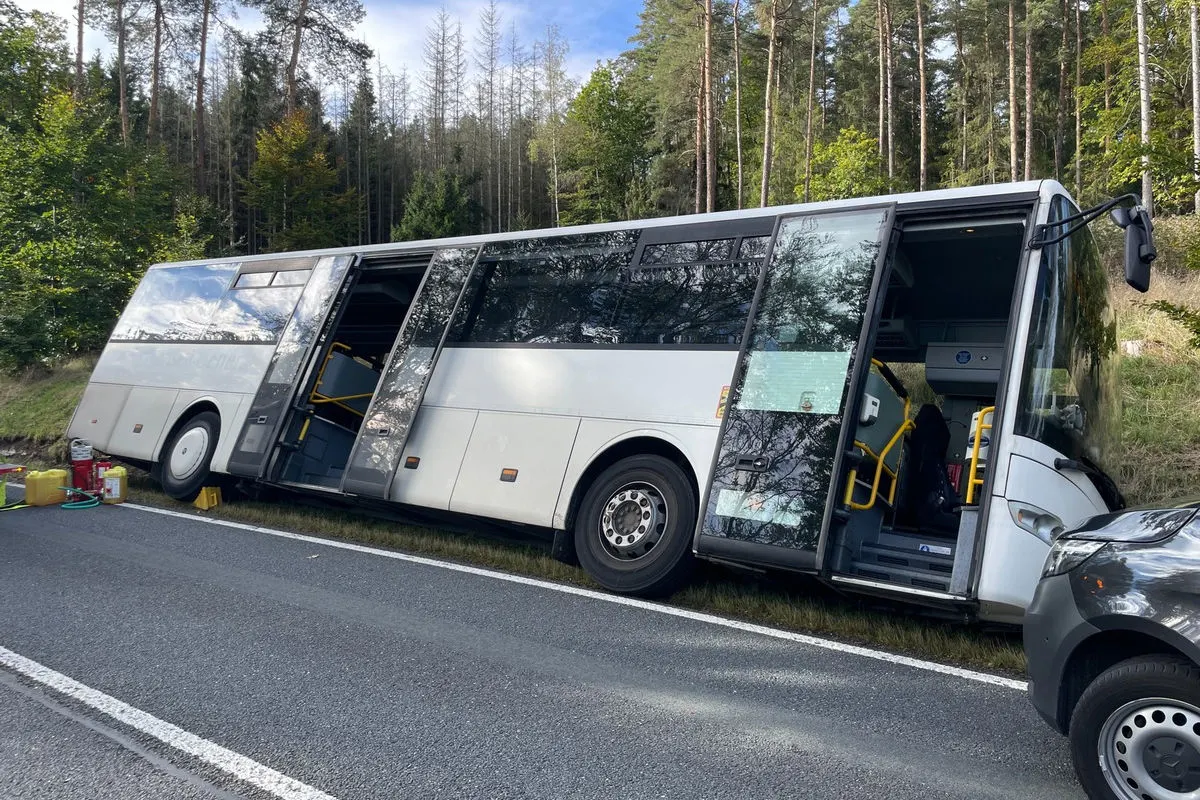 Ein leerer Linienbus steht schräg am Straßenrand einer Landstraße. Seine Türen sind geöffnet, und die Umgebung zeigt einen Wald im Hintergrund.