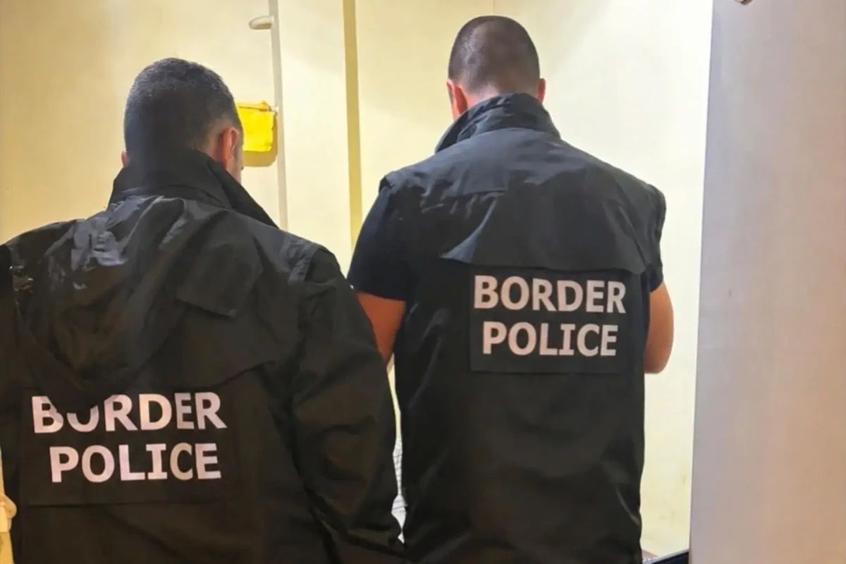 Zwei Männer in Jacken mit der Aufschrift 'BORDER POLICE' stehen mit dem Rücken zur Kamera in einem hell erleuchteten Raum.
