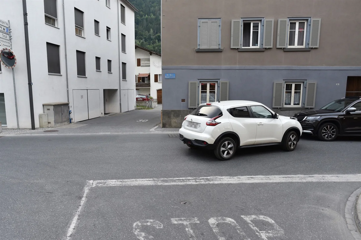 Ein weißes Auto steht an einer städtischen Kreuzung. Mehrfamilienhäuser umringen die Straßenecke.