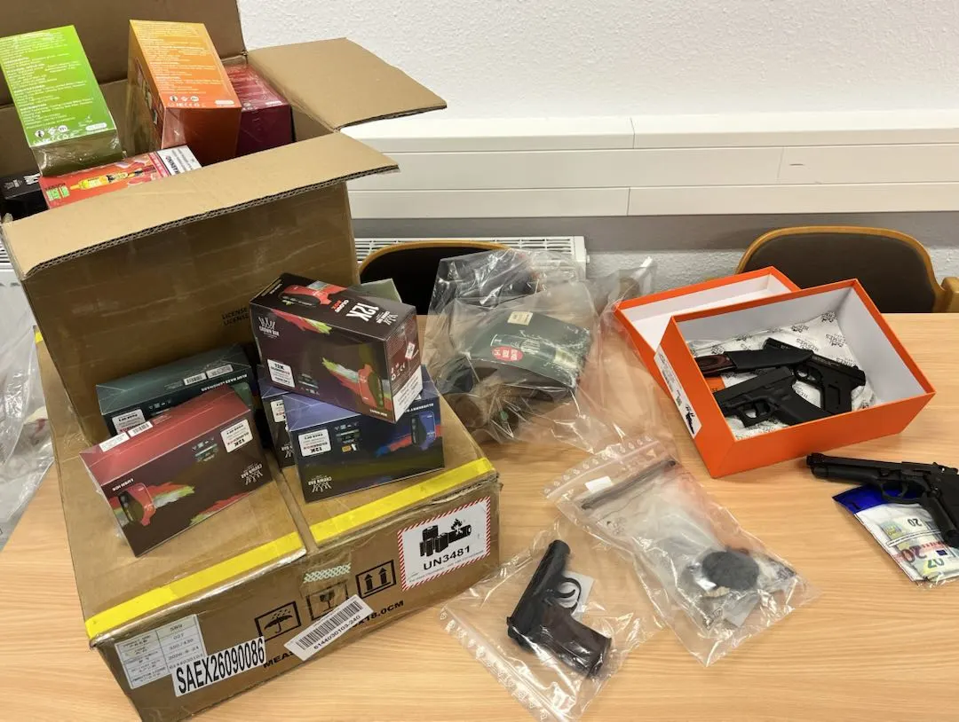Auf einem Tisch liegen geöffnete Kartons mit Verpackungen und mehrere Waffen, die in Plastikbeuteln verpackt sind, als Beweismaterial gesichert.
