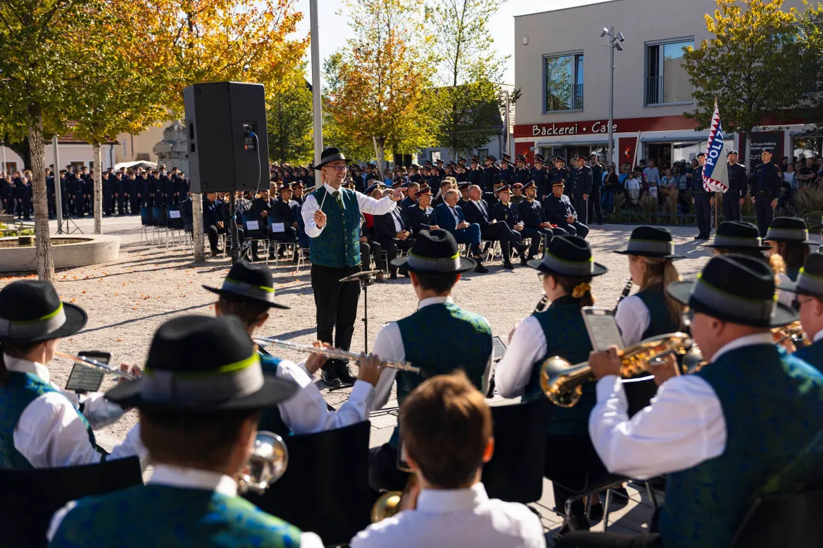 Ein Dirigent steht vor einer Musikkapelle, die bei einer Veranstaltung im Freien spielt. Im Hintergrund sitzen Polizisten in Uniform.