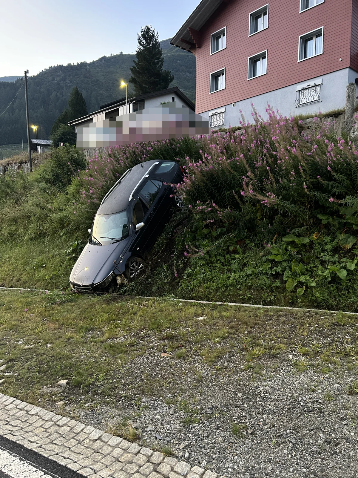 Ein Auto ist eine Böschung hinuntergerutscht und steckt in einem Hang fest. Es befindet sich vor einem großen Wohnhaus in einer ländlichen Gegend.