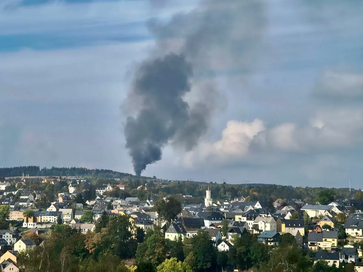 Eine größere Menge schwarzer Rauch steigt vom Horizont über einer städtischen Landschaft mit zahlreichen Gebäuden empor. Der Himmel ist leicht bewölkt, es ist tagsüber.