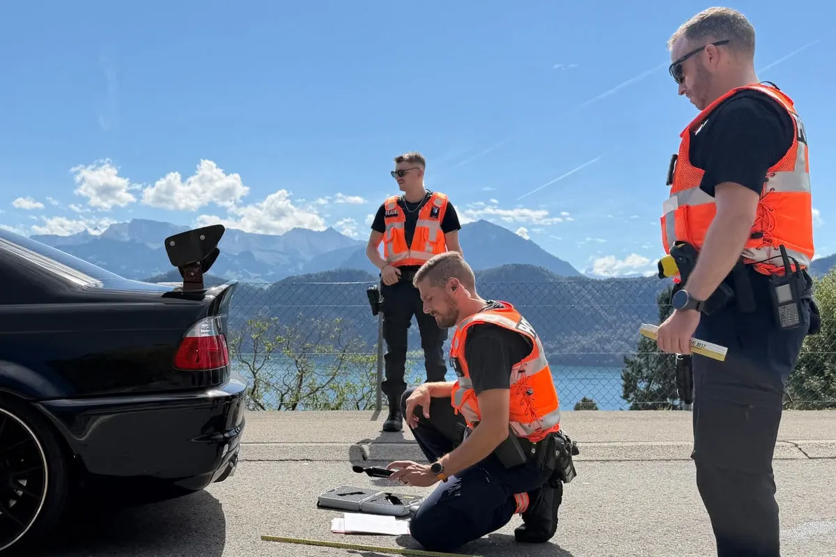 Drei Polizisten in Warnwesten stehen und knien auf einer Straße neben einem Pkw, vor einer Berg- und Seelandschaft.