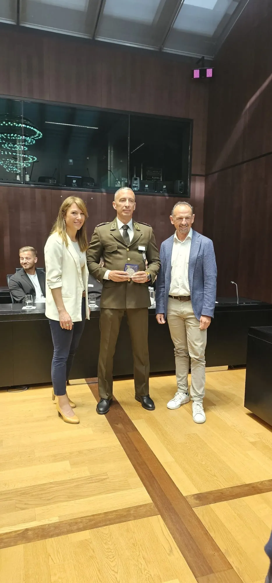 In einem modernen Konferenzraum posieren drei Personen für ein Foto. Die Person in der Mitte trägt eine Uniform und hält eine Medaille in den Händen.