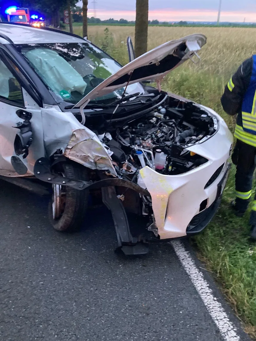 Ein stark beschädigtes weißes Auto steht an einer Landstraße. Die Front des Autos ist eingedrückt, und die Airbags sind ausgelöst.