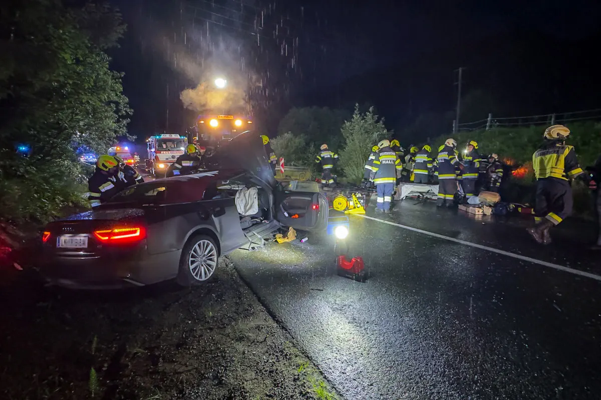 Ein beschädigtes Auto steht auf einer nassen Straße, umgeben von Feuerwehrleuten bei Nacht. Mehrere Rettungskräfte sind im Einsatz.