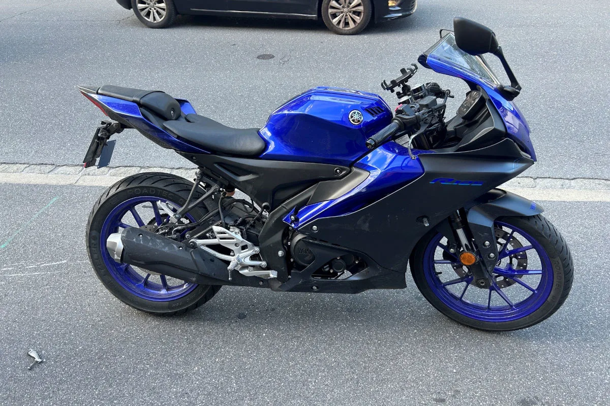 Ein blaues Motorrad ist seitlich auf einer Asphaltstraße geparkt. Im Hintergrund ist ein Teil eines schwarzen Autos zu sehen.
