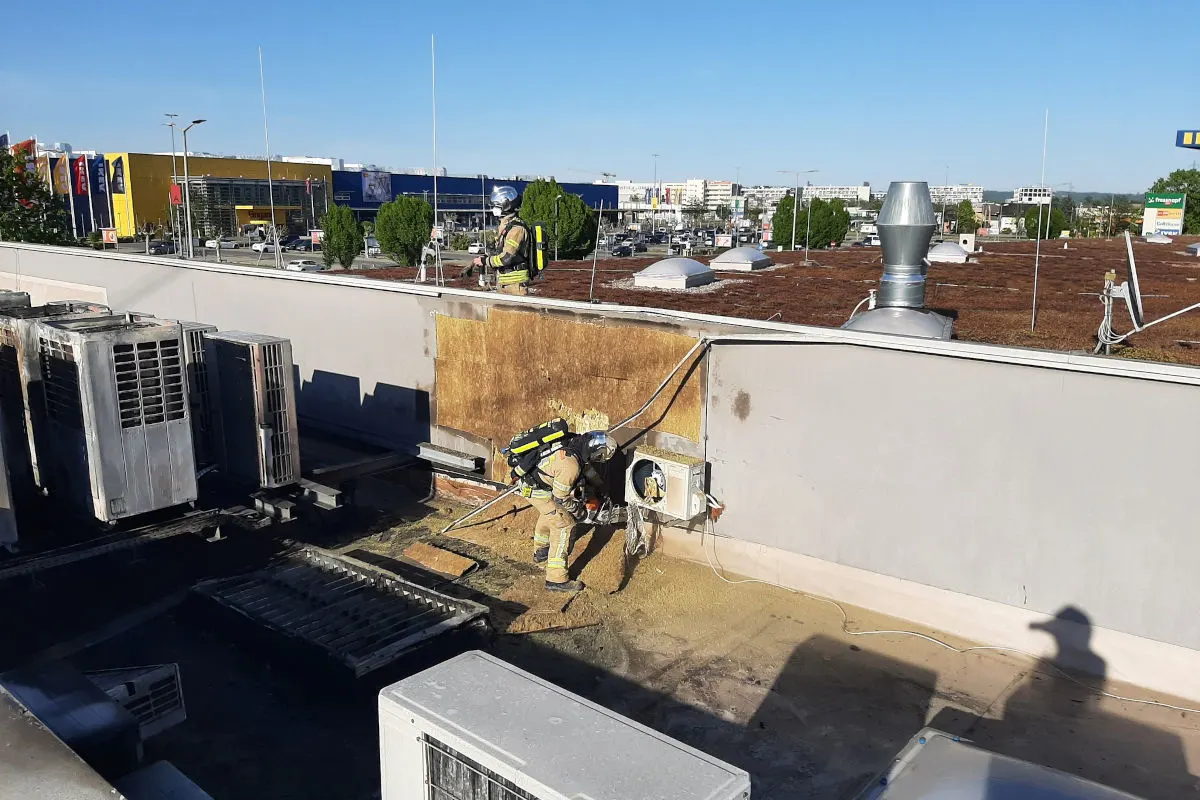Zwei Feuerwehrleute sind auf einem Dach zu sehen. Sie tragen Schutzanzüge und arbeiten an einer Fassade.