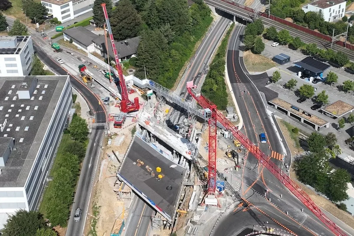 Luftaufnahme einer Baustelle mit zwei großen roten Kränen, umgeben von Straßen und Baugeräten. Die Baustelle ist an einer Kreuzung gelegen.