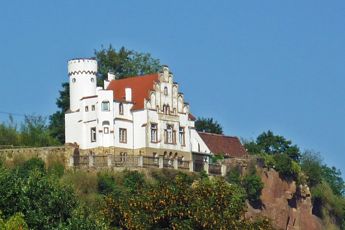 Ein weißes Gebäude mit einem roten Dach steht auf einem bewaldeten Hügel. Der Himmel ist klar und blau.