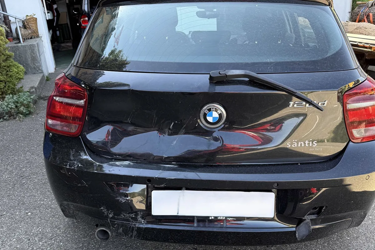 Die Aufnahme zeigt den beschädigten Kofferraum eines schwarzen BMWs mit sichtbaren Dellen und Kratzern. Das Fahrzeug steht auf einer asphaltierten Fläche vor einer Garage.