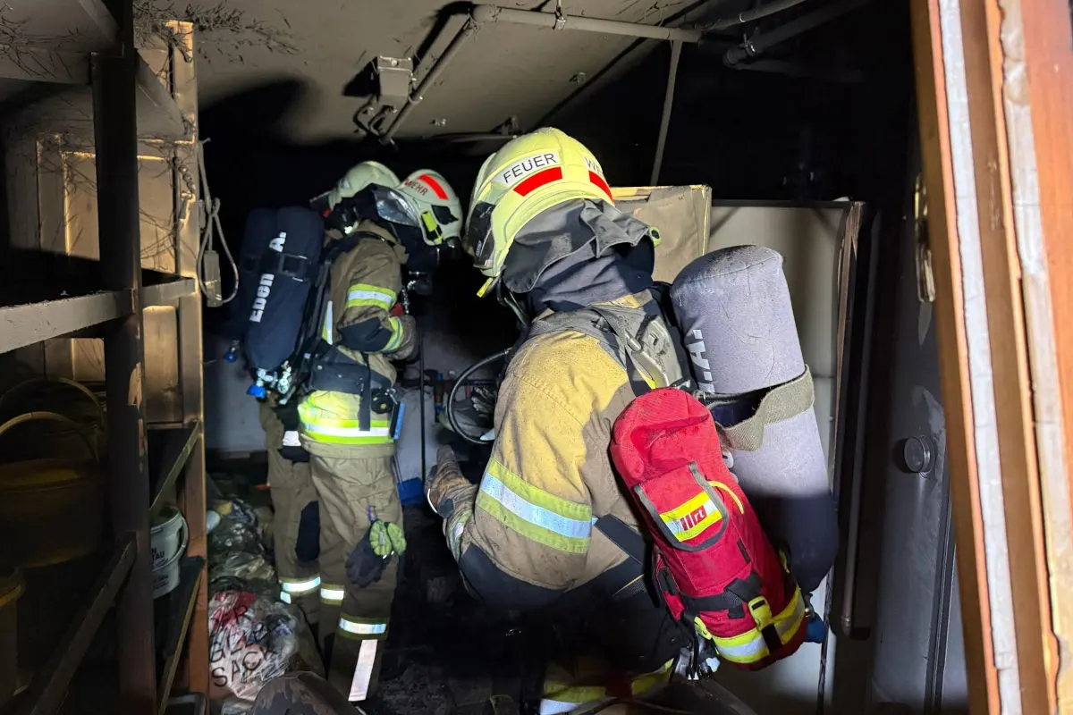 Zwei Feuerwehrleute mit Atemschutzmaske und Einsatzkleidung durchsuchen einen dunklen, engen Raum mit verrußten Wänden.