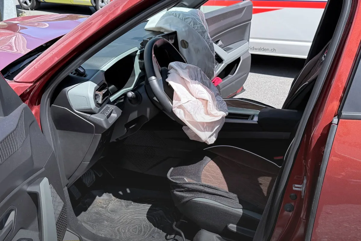 Ein ausgelöster Airbag ist im roten Auto sichtbar, die Fahrertür steht offen. Ein Ambulanzfahrzeug ist im Hintergrund erkennbar.