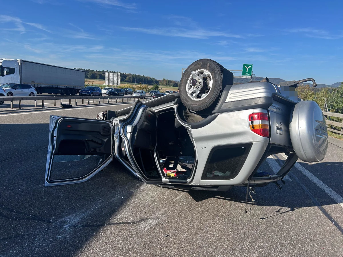 Ein silbernes Auto liegt kopfüber auf einer Autobahn. Im Hintergrund sind weitere Fahrzeuge und ein Lastwagen zu sehen. Der Unfall scheint unter klarem Himmel bei Tageslicht geschehen zu sein.